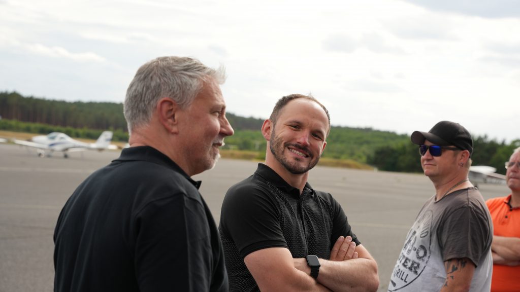 Gruppe von Männern am Flugplatz, lächelnder Mann mit verschränkten Armen im Vordergrund, Flugzeuge und grüne Landschaft im Hintergrund.