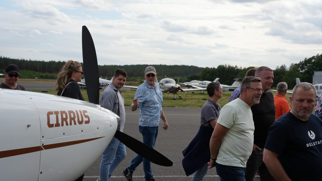 Gruppe von Menschen vor einem Cirrus-Flugzeug auf einem Flugplatz, mehrere Kleinflugzeuge im Hintergrund und lockere Stimmung beim Rundgang.